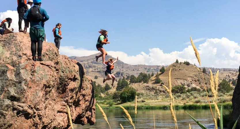 two outward bound students wearing life jackets jump off of a rocky overlook into water while others look on